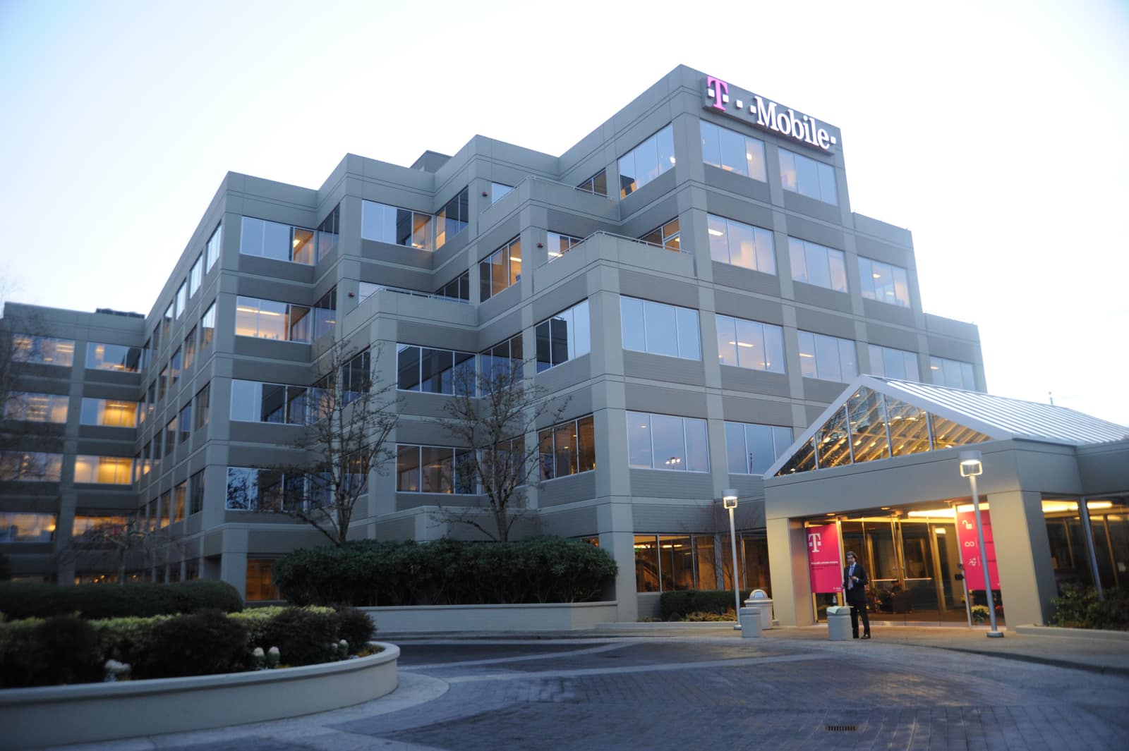 T-Mobile's corporate headquarters in the Factoria district of Bellevue, Washington, photographed at dusk. The magenta T-Mobile wordmark is lit on the upper facade of a grey stone-and-glass office block, office floors are lit behind the windows, and a single figure stands under the glazed entrance canopy.